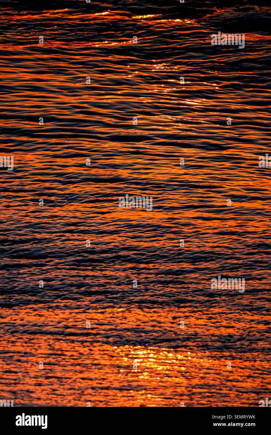 Leuchtend orange Sonnenaufgang reflektiert über ruhiges Meereswasser mit abstrakten Wellenstrukturen. Stockfoto