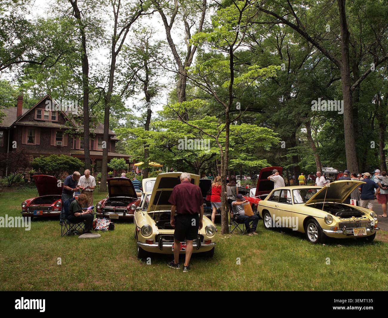Ein Treffen von MG-B und MG-B/GT auf einer lokalen All British Car Show. Leute sahen, wie sie die verschiedenen Autos in einem schattigen Waldgebiet mit einem alten Haus durchschauten. Stockfoto
