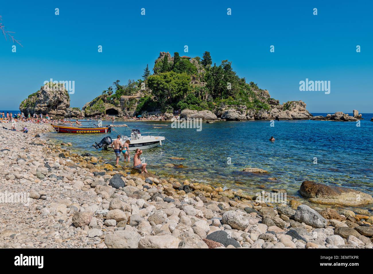 Besucher waten und entspannen sich in der Nähe kleiner Motorboote an der felsigen Kieselküste von Isola Bella, einem kleinen, von Bäumen bewachsenen Inselnaturschutzgebiet gleich neben der Küste Stockfoto