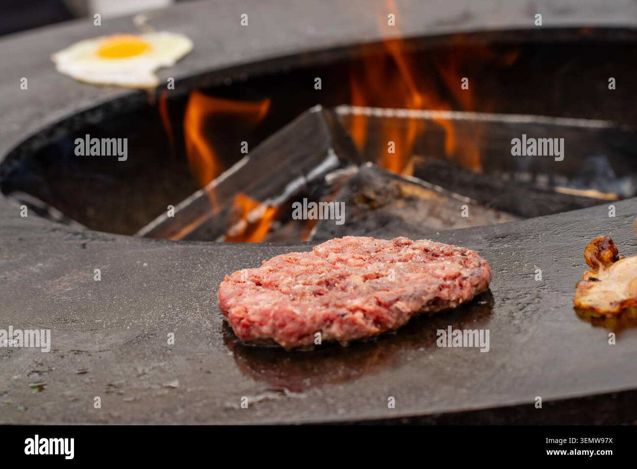 Frisches Fleischschnitzel und ein ganzes Ei-Braten auf einer runden Grillstation aus Metall, wobei brennende Holzscheite Rauch während des Cateringservice im Freien ausstoßen Stockfoto