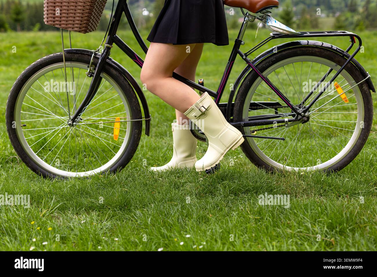 Vintage-Fahrradräder Und Regenstiefel. Nahaufnahme einer Frau in cremefarbenen Regenstiefeln, die neben einem alten Fahrrad auf frischem grünem Gras steht. Stockfoto