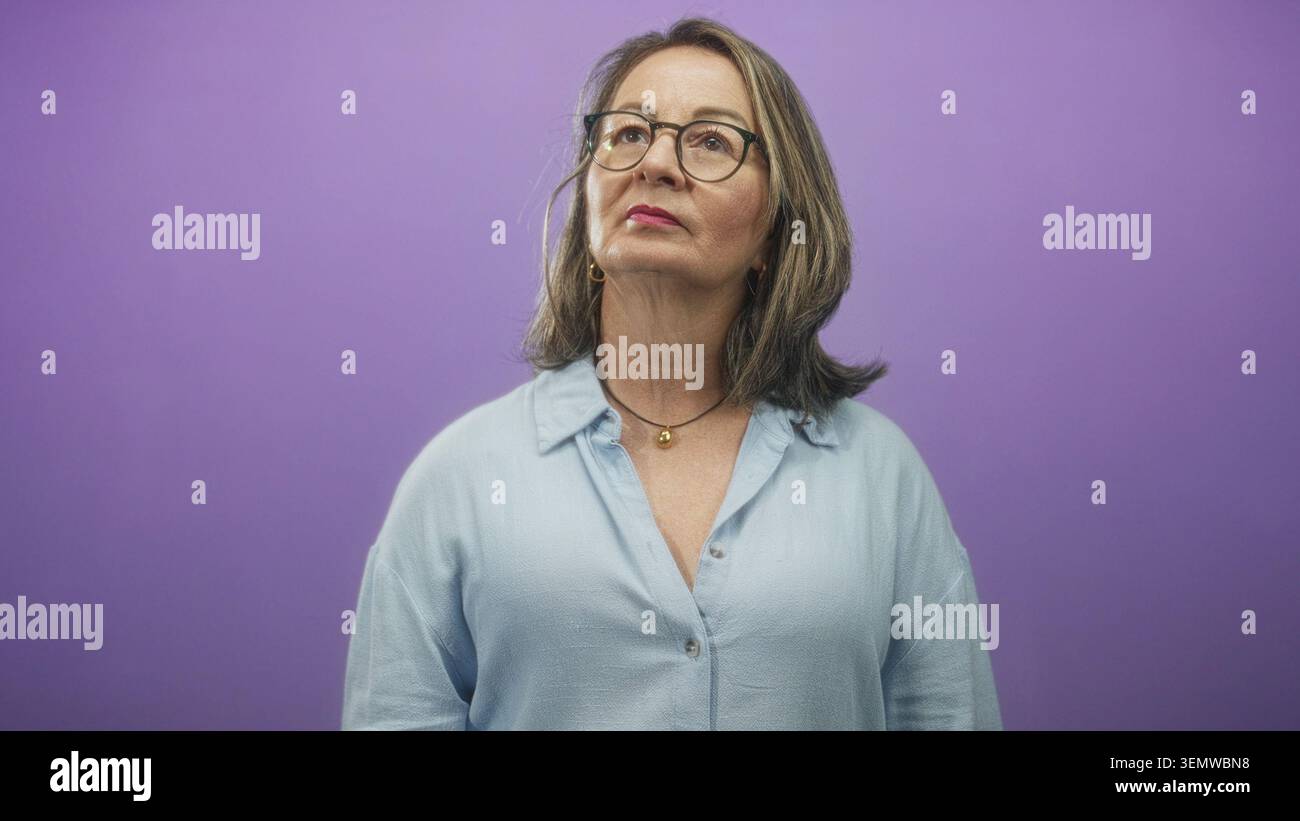 Frau hispanic Senior mit grauem Haar und Brille zeigt den Zeigefinger nach oben, während sie vor einem violetten Studio-Hintergrund aufblickt; Neugier. Stockfoto