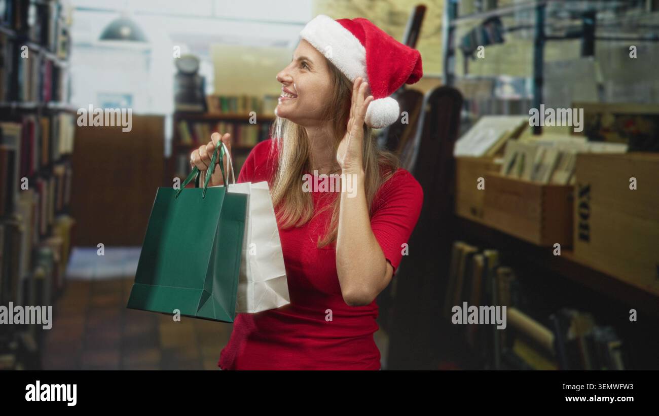 Frau im weihnachtsmann-Hut mit grünen und weißen Einkaufstaschen, lächelnd und Tassen Ohr, während sie in der Buchhandlung aufblickt; Urlaubsfreude. Stockfoto