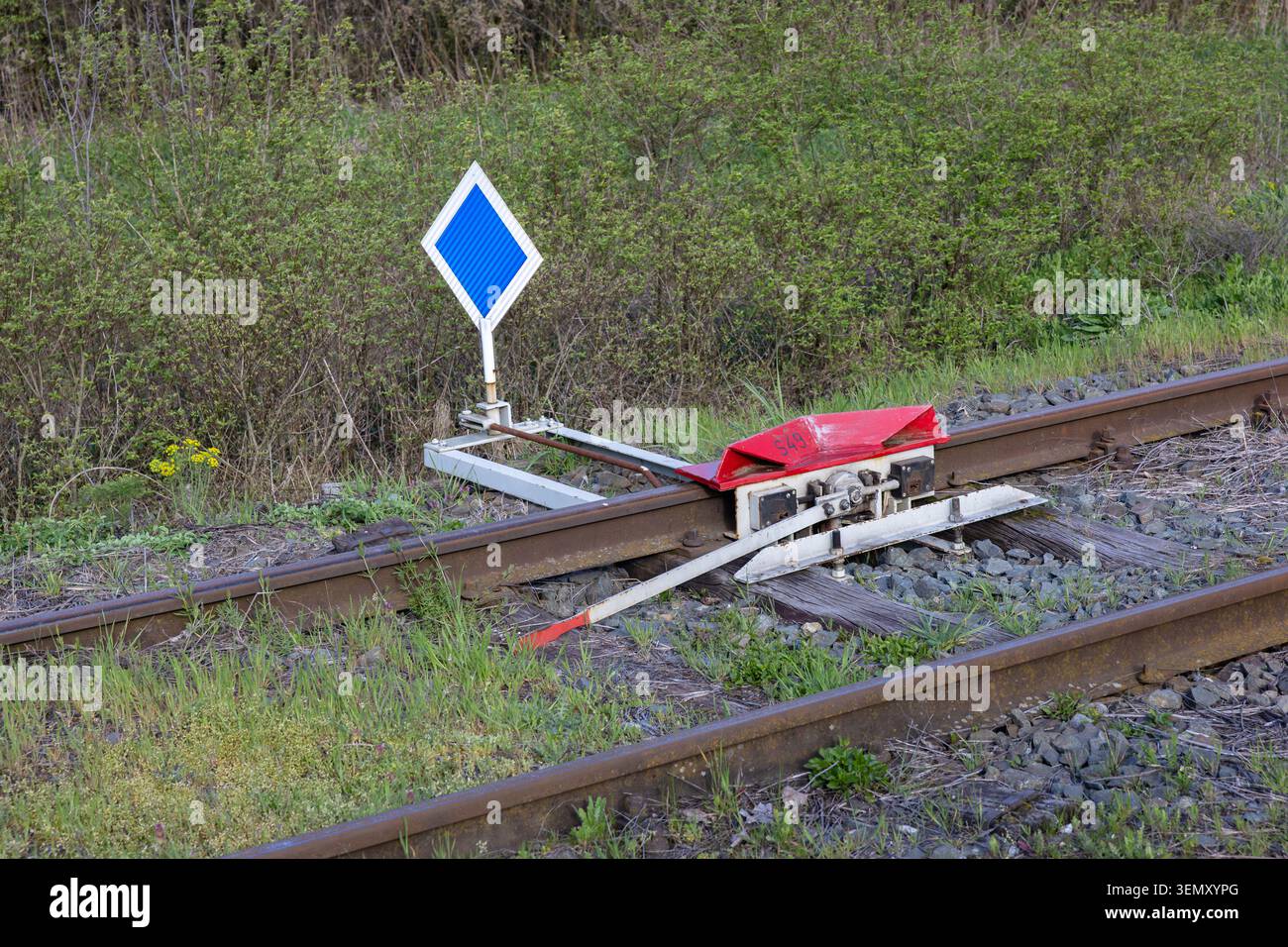 Ein Bahnschalter und eine rote Bahnhaltestelle neben einem blauen Diamantschild auf rostigen Schienen, umgeben von grünem Gras und Sträuchern. Die ländliche Eisenbahninfrastruktur ruft hervor Stockfoto