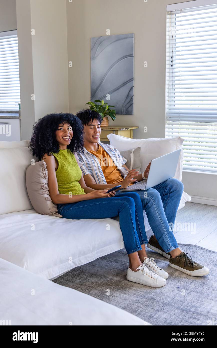 Ein Paar Afroamerikanerin, ein asiatischer Mann, der im Wohnzimmer auf dem Sofa sitzt, Laptop und Fernbedienung hält Stockfoto