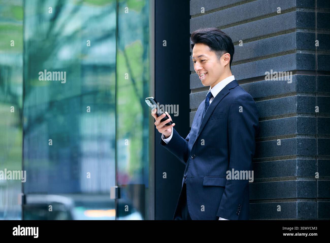 Ein lächelnder japanischer Geschäftsmann in seinen 40ern hält ein Smartphone, das draußen an eine Wand gelehnt ist Stockfoto