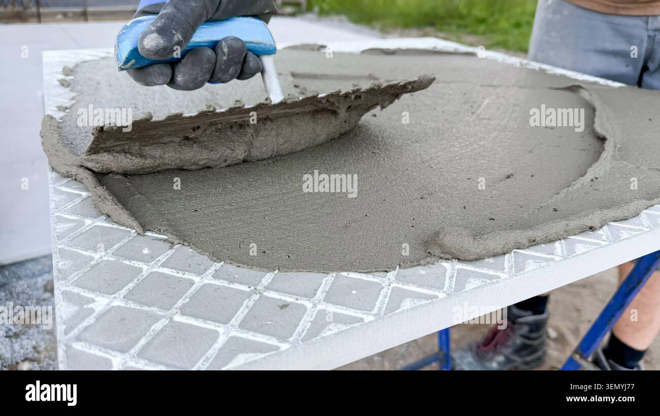 Arbeiter, der mit einer Kelle graue Zementmischung auf eine strukturierte Fliesenoberfläche aufträgt, mit einem verschwommenen grünen Hintergrund, der auf eine Baustelle im Freien hinweist Stockfoto