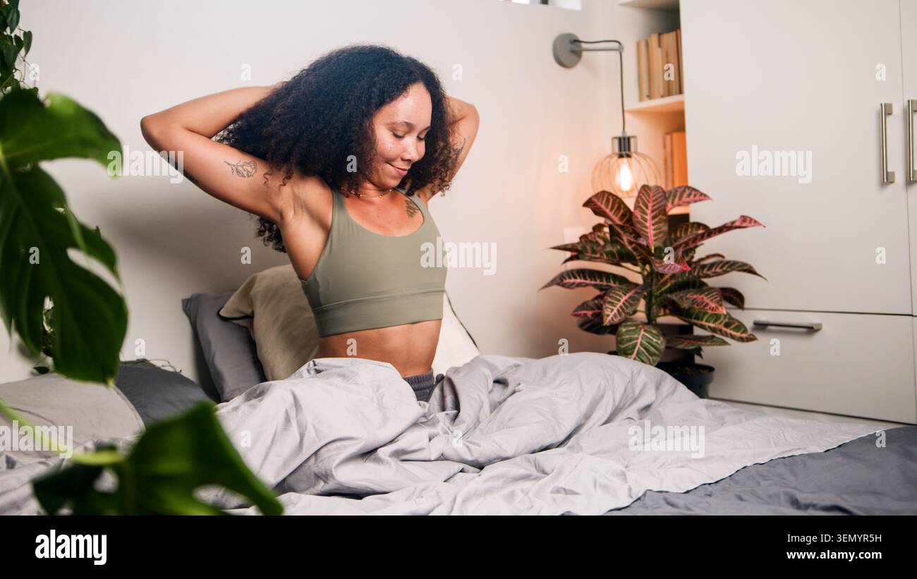 Junge Frau dehnt sich im Bett während der entspannten Morgenroutine im gemütlichen, mit Pflanzen gefüllten Schlafzimmer Stockfoto