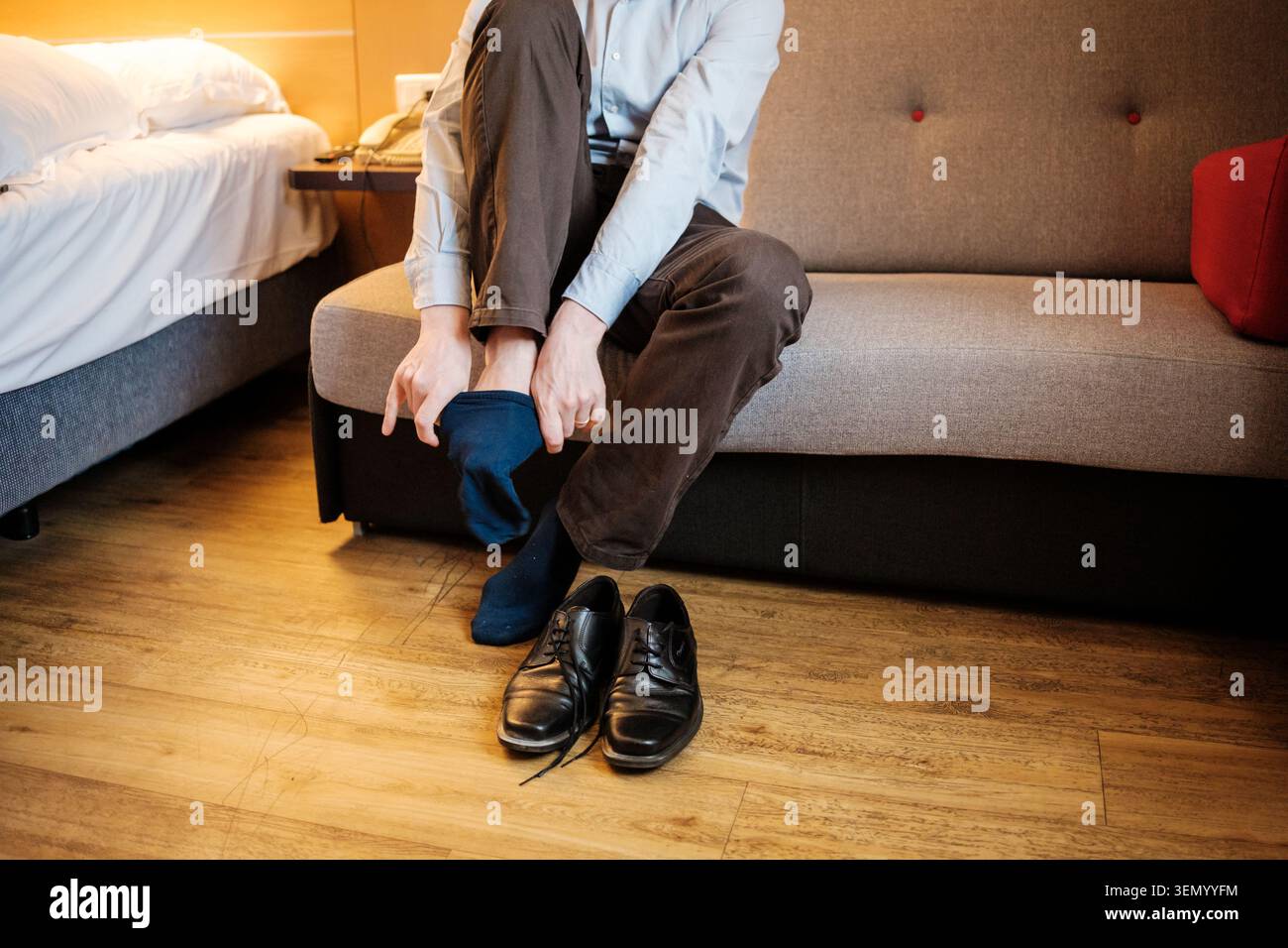 Mann sitzt auf einer Couch und zieht blaue Socken neben seinen Schuhen in einem Hotelzimmer an Stockfoto
