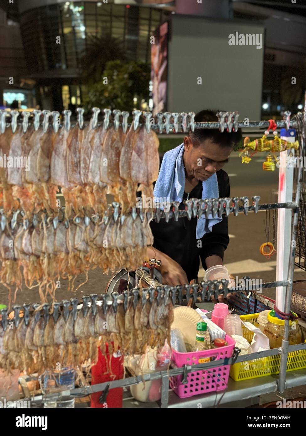 Reihen von getrocknetem Tintenfisch hängen wie Vorhänge über einem Nachtmarkt-Grill, während ein Koch die nächste Straßenfood-Bestellung zubereitet. - Smartphone-aufgenommenes Stockfoto