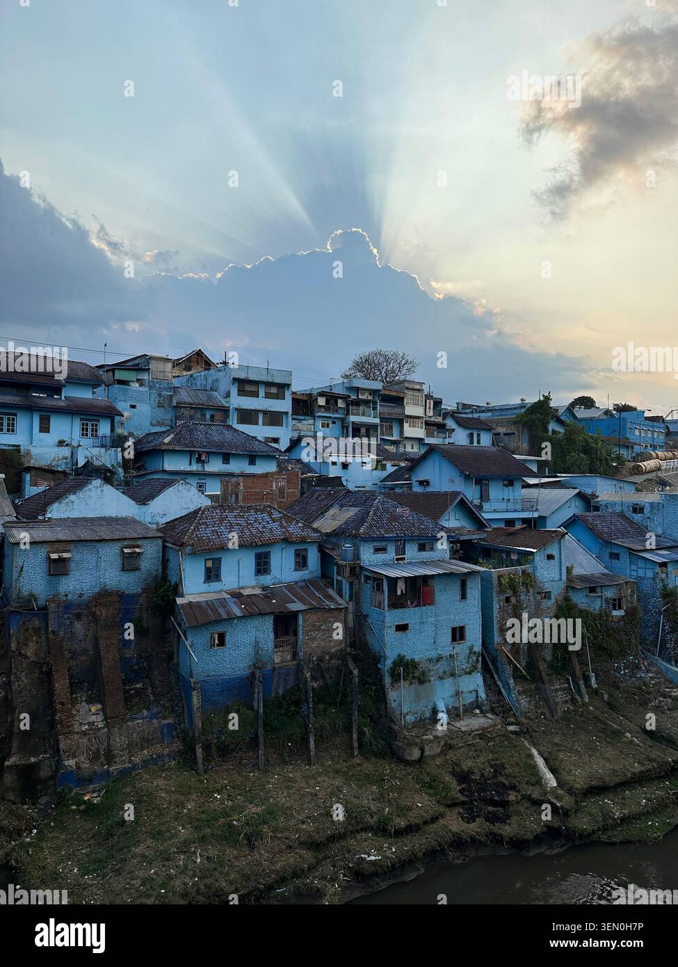 Sich blau fühlen: Eine Nachbarschaft, die in Blautönen unter strahlenden Sonnenstrahlen das Flussufer säumt, Kampung Biru Arema, Malang, Indonesien - Smartphone-aufgenommenes Stockfoto