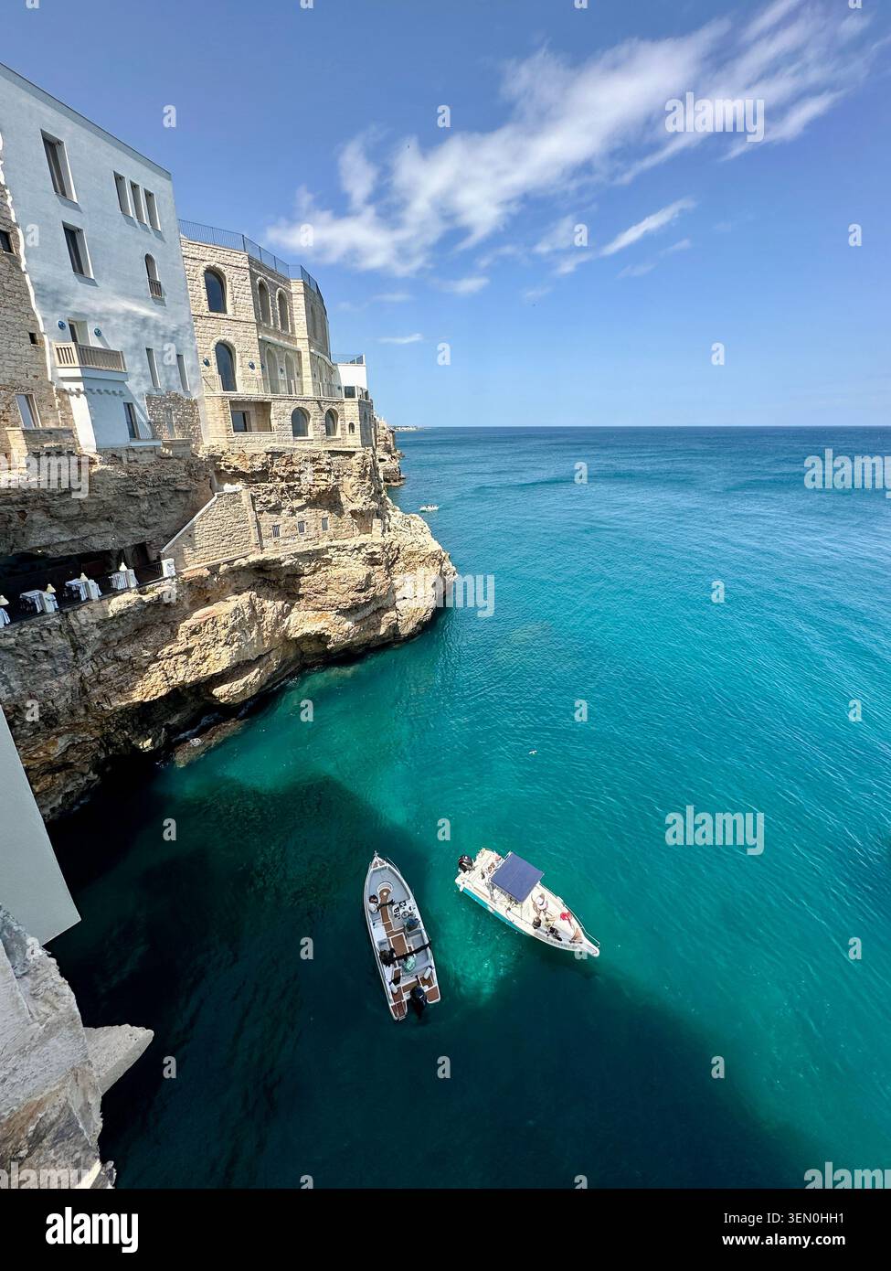 Weiße Klippengebäude treffen auf strahlend türkisfarbenes Wasser, während zwei kleine Boote unterhalb einer sonnenbeschienenen Küstenstadt treiben - Smartphone-aufgenommenes Stockfoto