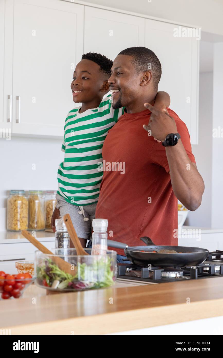 Afroamerikanischer Vater, der Sohn hält, bereitet auf der Kücheninsel Essen mit einer Pfanne vor Stockfoto