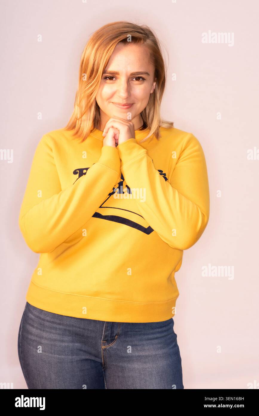 Hannah Witton steht in einem Studio mit neutralem Hintergrund. Sie lächelt und schließt ihre Hände zusammen. Sie trägt ein gelbes Sweatshirt und Jeans. Stockfoto