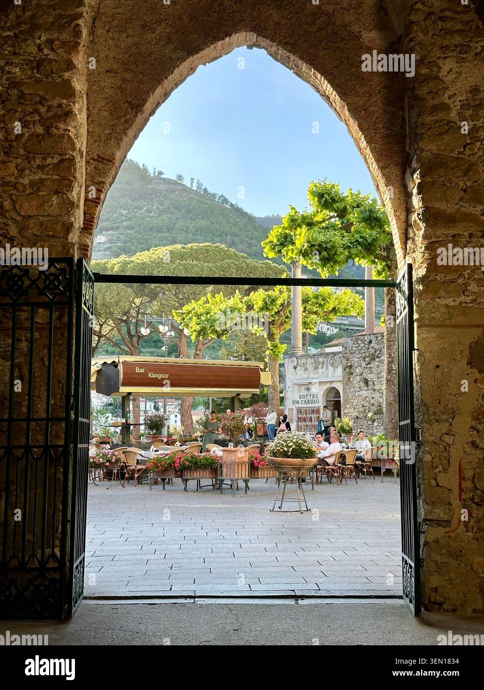 Ein spitzer Steinbogen öffnet sich zu einem sonnenbeschienenen Dorfplatz und umrahmt Cafétische, Blumen und grüne Hügel dahinter - Smartphone-aufgenommenes Stockfoto