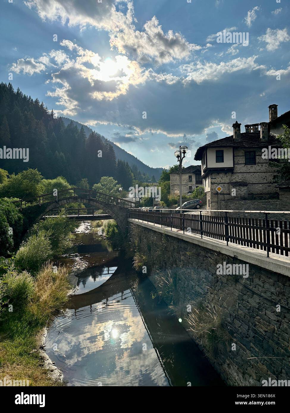 Sonnenlicht bricht durch Bergwolken über einem spiegelglatten Dorfkanal und verwandelt das Tal in flüssiges Gold. - Smartphone-aufgenommenes Stockfoto
