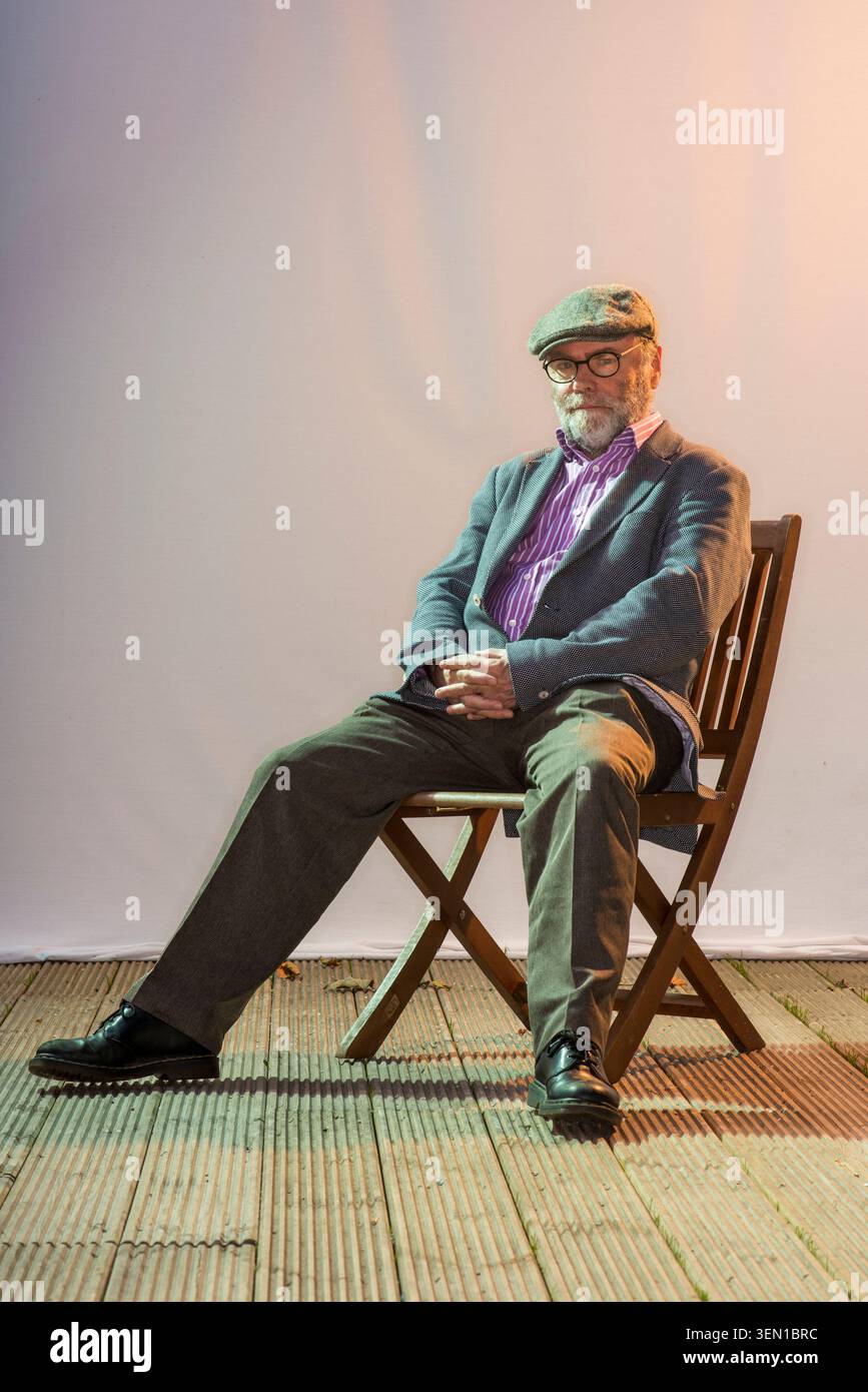 Malachi O'Doherty sitzt drinnen immer noch auf einem Holzstuhl. Er trägt eine Brille und eine flache Kappe, während das Licht hinter ihm eine warme Atmosphäre erzeugt. Der Spac Stockfoto