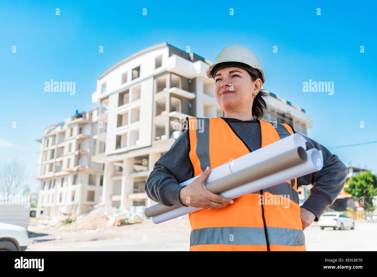 Eine junge hispanische Frau mit Schutzhelm und orangefarbener Sicherheitsweste hält auf einer Baustelle mit Gebäuden im Hintergrund Blaupausen. Stockfoto
