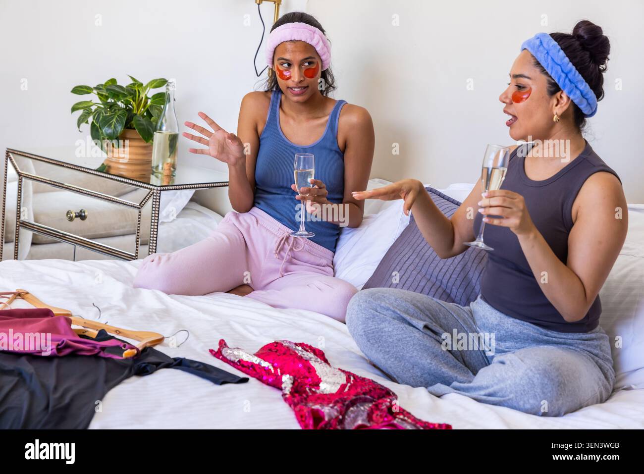 Verschiedene Freundinnen plaudern und trinken Champagner auf dem Bett zu Hause mit Paillettenkleid in der Nähe Stockfoto