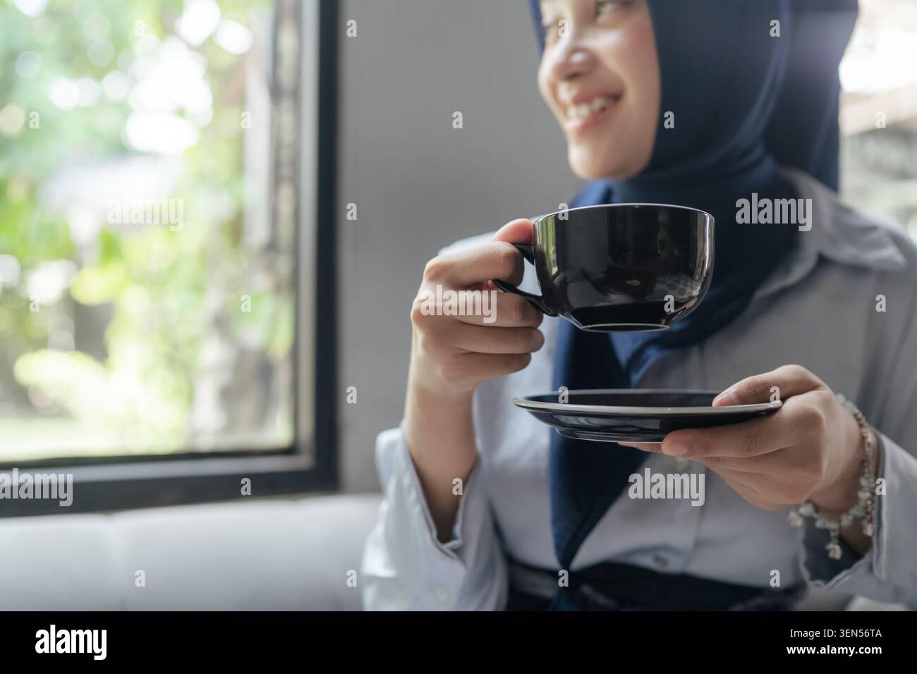 Lächelnde Frau im Hijab, die eine Tasse und Untertasse in der Nähe eines Fensters in einem hellen Café hält Stockfoto