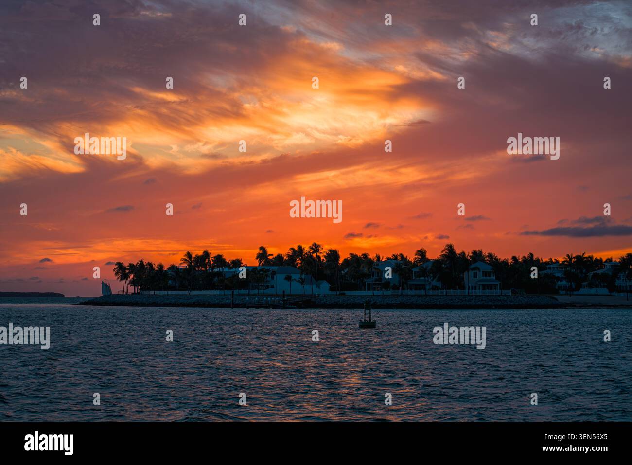 Das kleine Passagiersegelboot überquert das dunkle Ozeanwasser in der Nähe der Silhouette der Key West Island unter den roten Wolkenformationen während der Dämmerung. Stockfoto