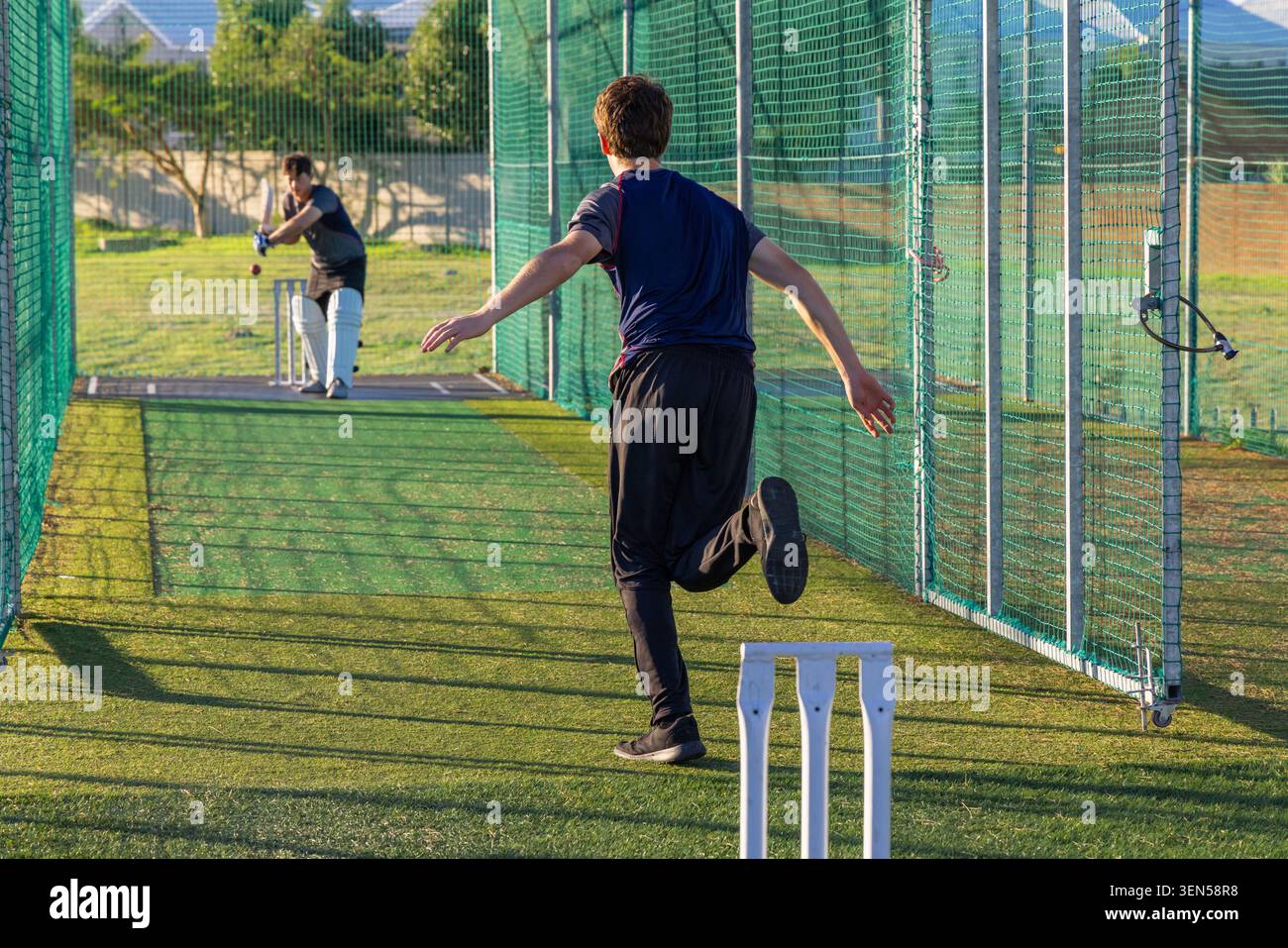 Zwei Männer üben Cricket im Netz auf Rasen, tragen Sportbekleidung, halten Fledermaus, Ball und weiße Stümpfe Stockfoto