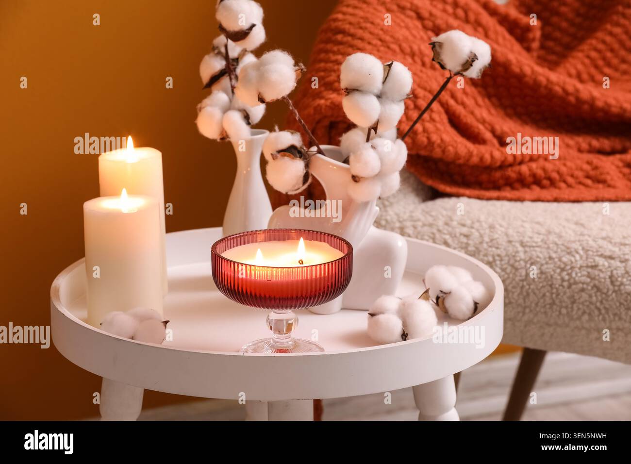 Gemütliches Ambiente mit zündeten Kerzen, Baumwollstielen und warmer Strickdecke auf einem Beistelltisch Stockfoto
