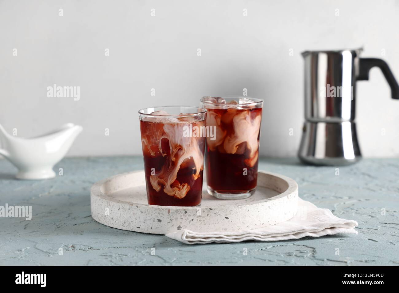 Zwei erfrischende Eiskaffee-Drinks mit Sahnewirbeln und Eiswürfeln auf einem Terrazzo-Tablett mit Mokakanne, eine weiße Leinenserviette wird unter die Gläser gestellt Stockfoto