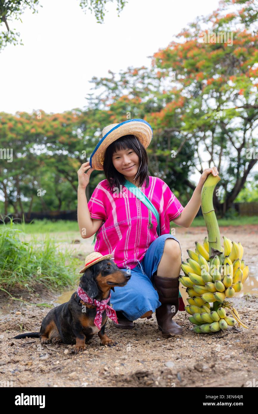 Frau Bananenhut rosa Kleid Garten Lächeln Junge Frau, die Bananenhut hält, während sie mit einem kleinen Hund im tropischen Garten sitzt und Strohhut und pi trägt Stockfoto