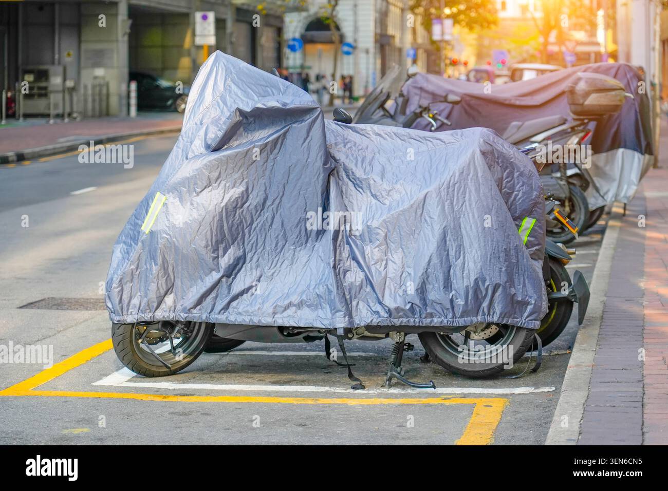 Das Motorrad ist mit einer grauen Schutzjacke bedeckt. Ein Roller mit Stoffschild steht auf dem Bürgersteig auf dem Stadtparkplatz. Wasserdichter Außenparkplatz Stockfoto