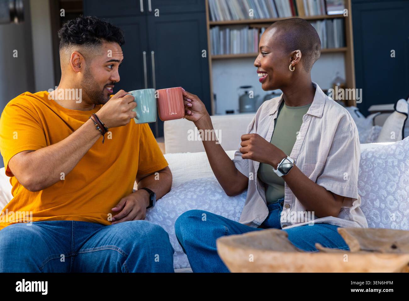 Diverse Paare sitzen auf dem Sofa zu Hause und knacken Keramikbecher, Mann orange T-Shirt, Frau beige Overshirt Stockfoto