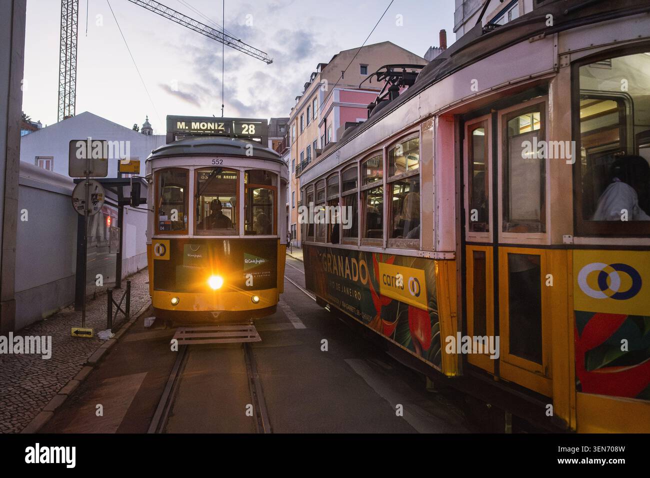 Zwei berühmte gelbe Straßenbahnen der Linie 28 fahren einander auf einer schmalen Kopfsteinpflasterstraße in Lissabon in der Abenddämmerung, Portugal. Die historischen Straßenbahnen Remodelado, ein Symbol Stockfoto