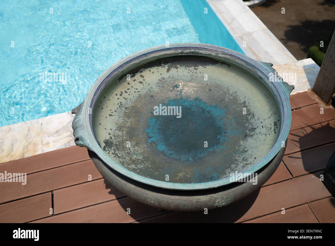 Eine verwitterte Bronzeschale mit grüner Patina hält Wasser auf einer Holzterrasse neben einem türkisfarbenen Pool in einem Luxusresort in der Nähe von Chiang Mai, Thailand Stockfoto