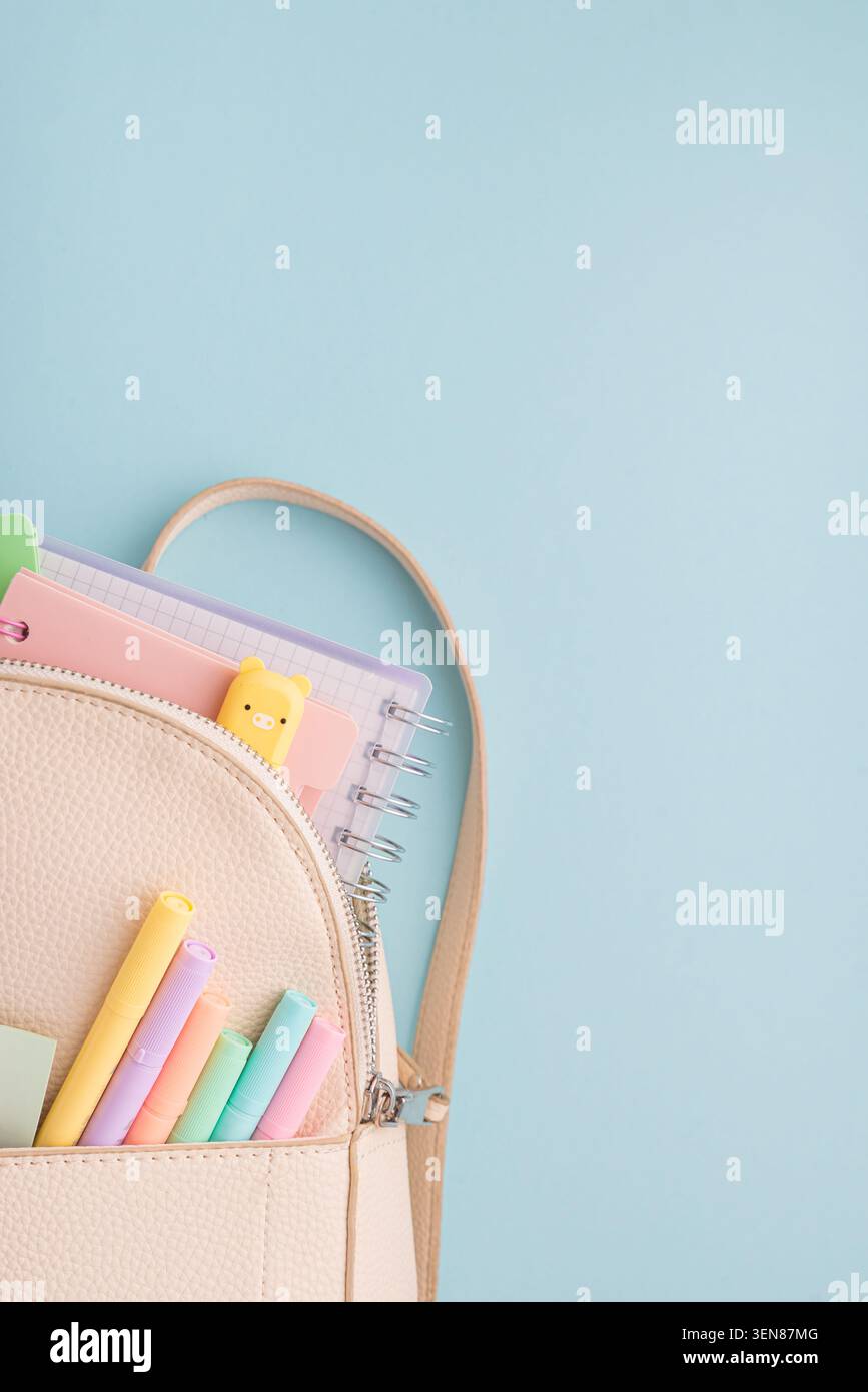 Der Rucksack aus beigefarbenem Kunstleder lässt sich öffnen, um spiralförmige Notizbücher, Textmarker in Pastellfarben, Stifte und ein niedliches Lesezeichen für Schweine auf weichem blauem Hintergrund zu entdecken Stockfoto