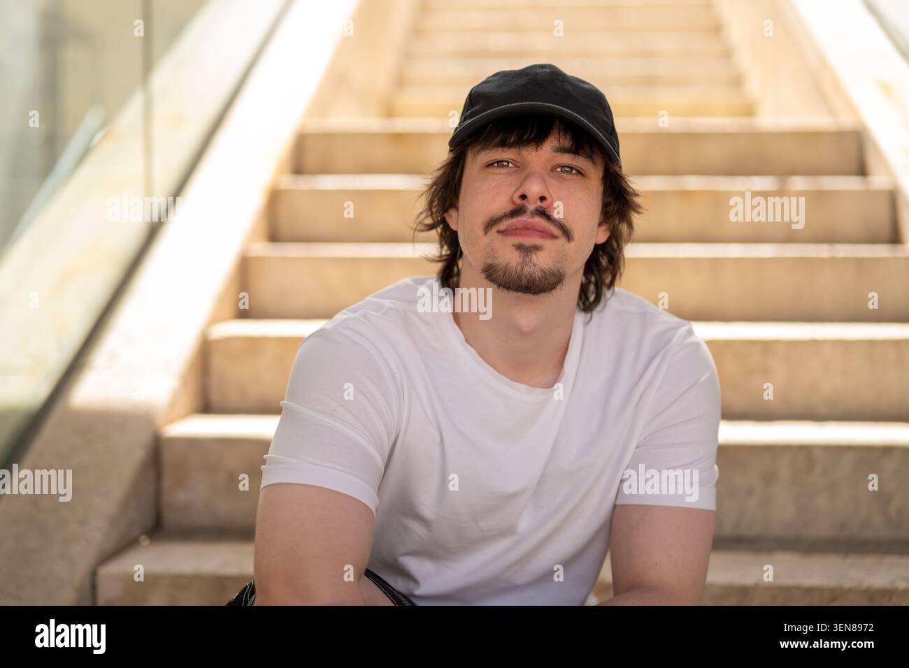 Junger 25-jähriger Mann, der auf der Treppe des modernen Komplexes sitzt. Stockfoto