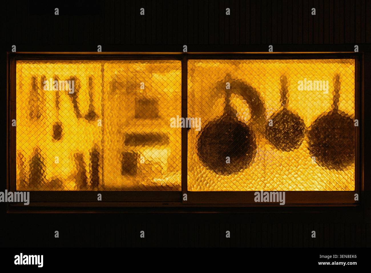 Warmes Licht leuchtet durch das Fenster der Küche des Restaurants und verwandelt Hängeutensilien in dunkle, vertraute Formen vor dem Glas, Tokio, Japan Stockfoto