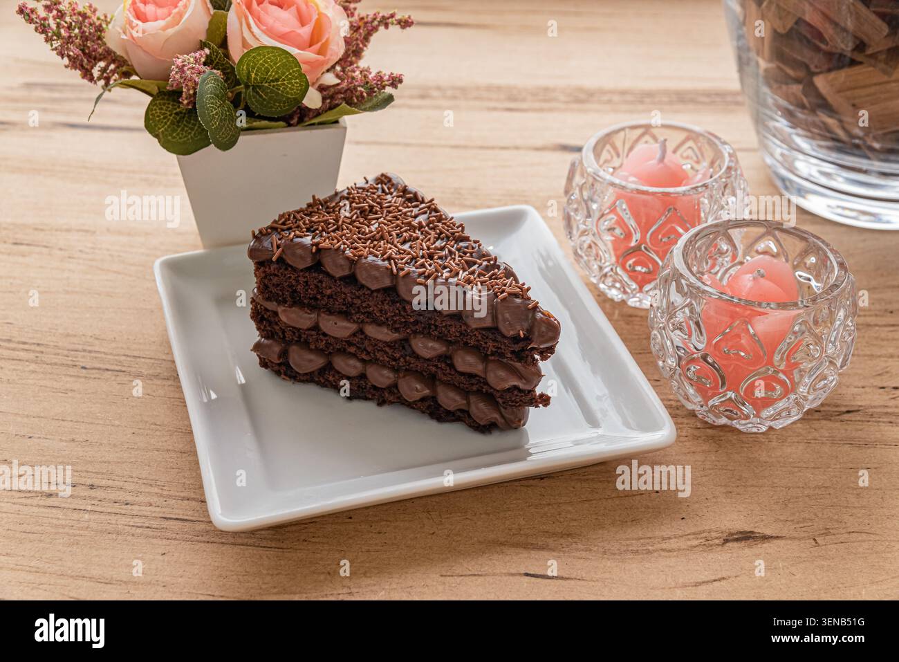 Kuchenscheibe auf weißem Teller, Kerzen und Blumenarrangement. Warme Studiobeleuchtung, die die reichhaltige, dunkle Schokolade hervorhebt, großartige Fotos von Speisen Stockfoto