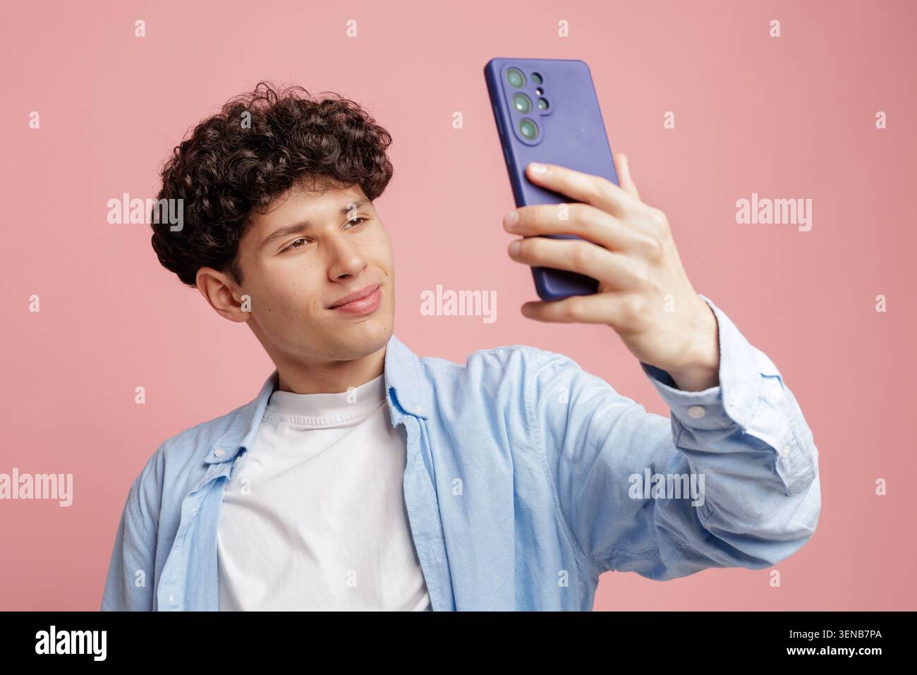 Ein junger Mann mit lockigen Haaren lächelt, während er mit seinem Smartphone ein Selfie macht und einen verspielten und modernen Lebensstil vor einem leuchtend rosa Backdro zeigt Stockfoto