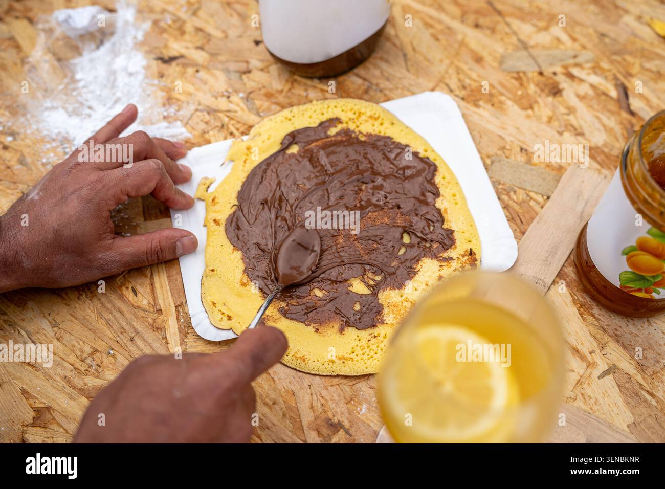 Die Hände der Person bereiten einen süßen Pfannkuchen zu, verteilen reichhaltige Schokoladen-Haselnusscreme auf der goldenen Oberfläche, mit einem erfrischenden Zitronengetränk und Marmeladenglas vor Stockfoto
