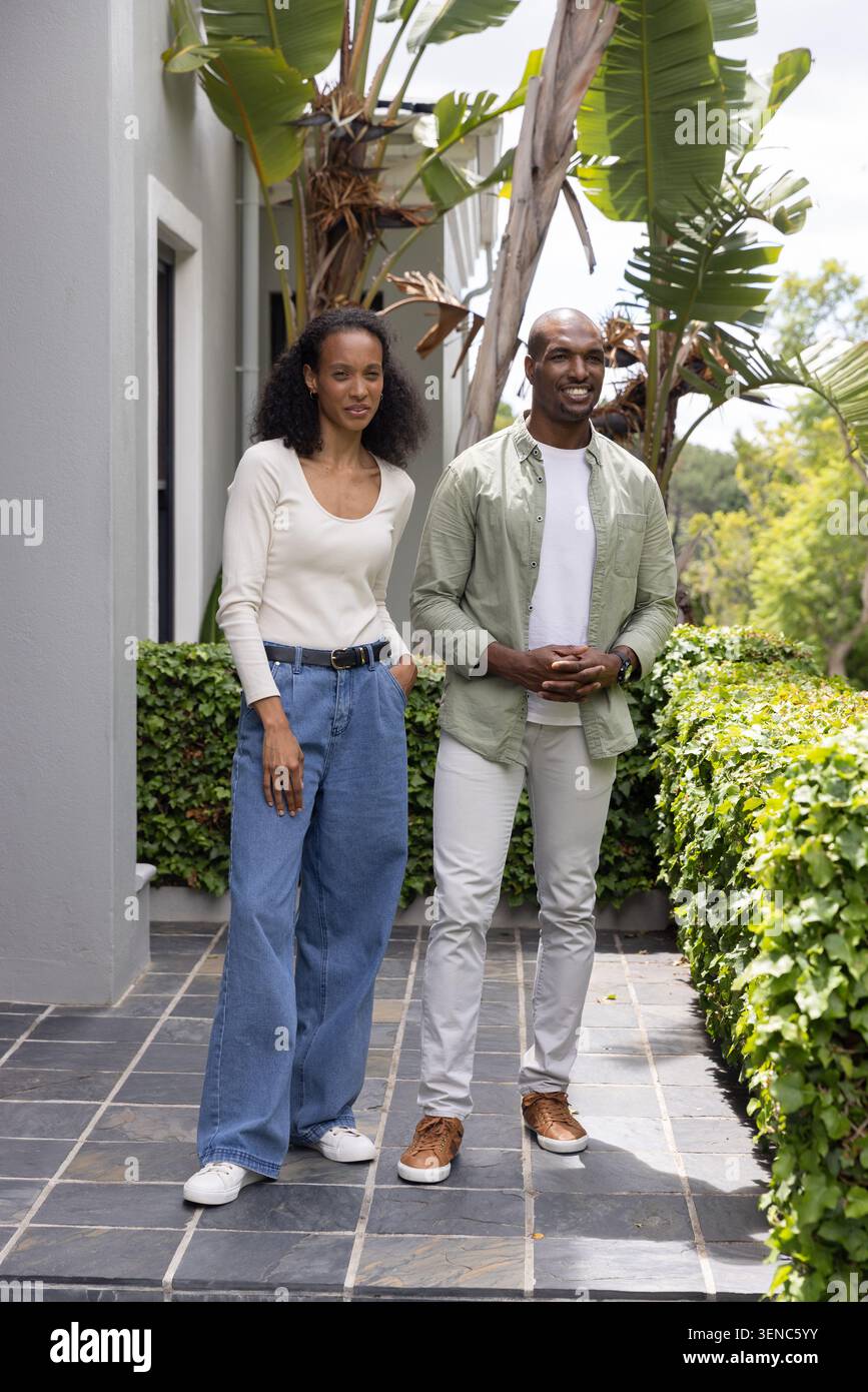 Paare posieren auf gekachelten Pfaden neben grauen Wänden, Hecken und Pflanzen, tragen Cremetop, Jeans, Turnschuhe Stockfoto