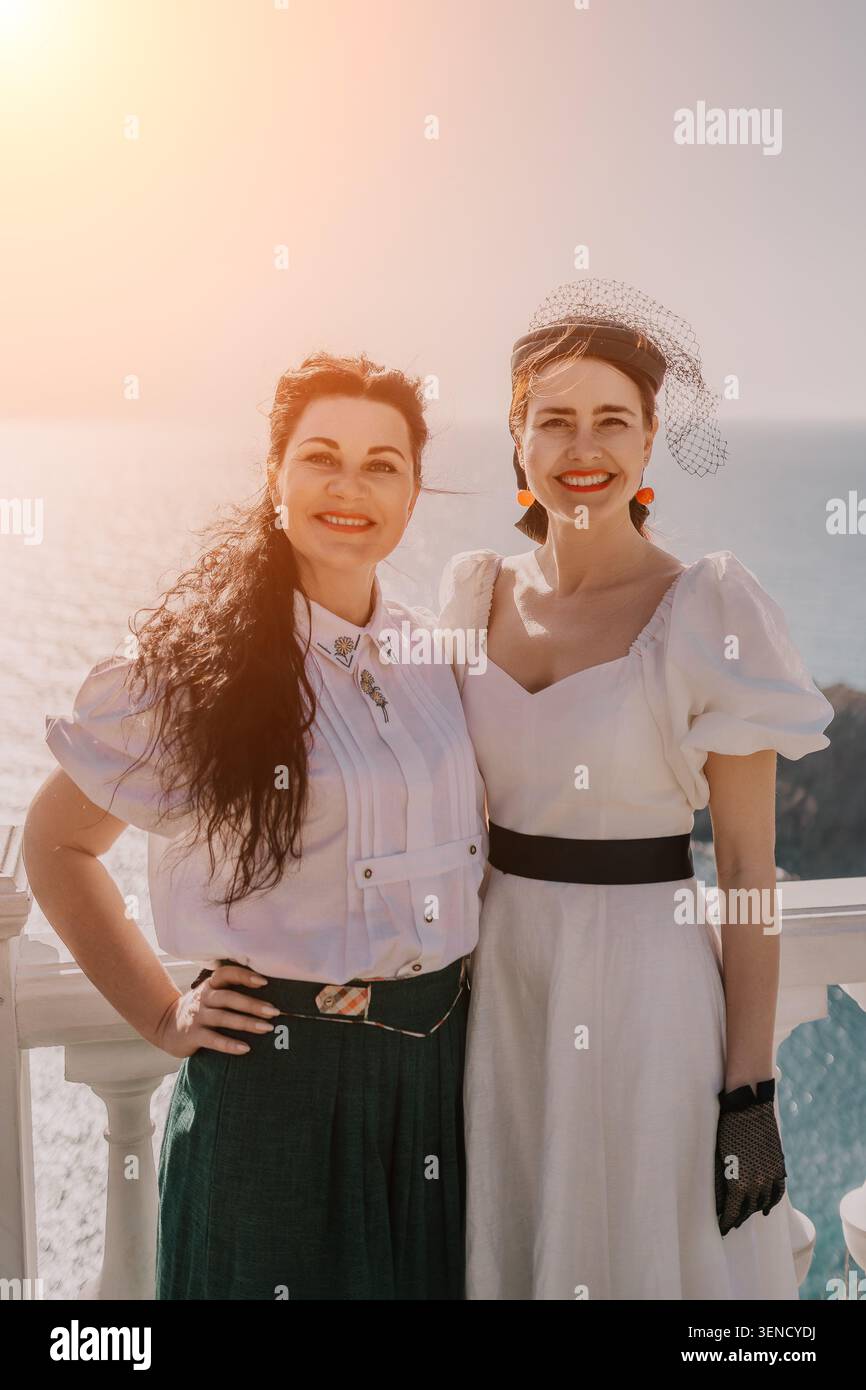 Freunde Frauen posieren im Urlaub auf einem sonnigen Balkon mit Blick auf das blaue mittelmeer in weißen Kleidern im Vintage-Stil Stockfoto