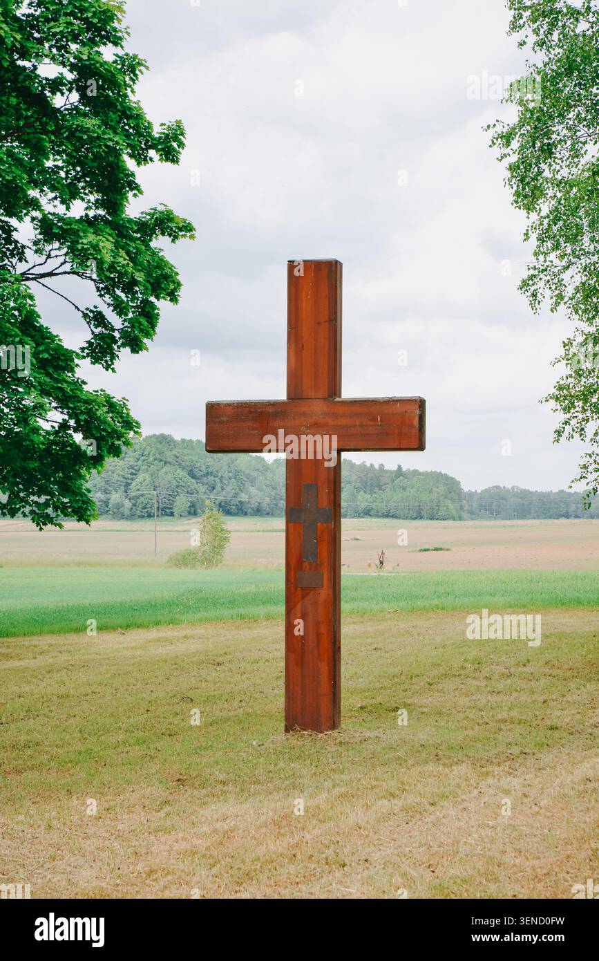 Riga, Lettland - 5. Juni 2026: Hölzernes christliches Kreuz auf einer friedlichen ländlichen Wiese, umgeben von Bäumen und offenem Ackerland unter bedecktem Himmel. S Stockfoto