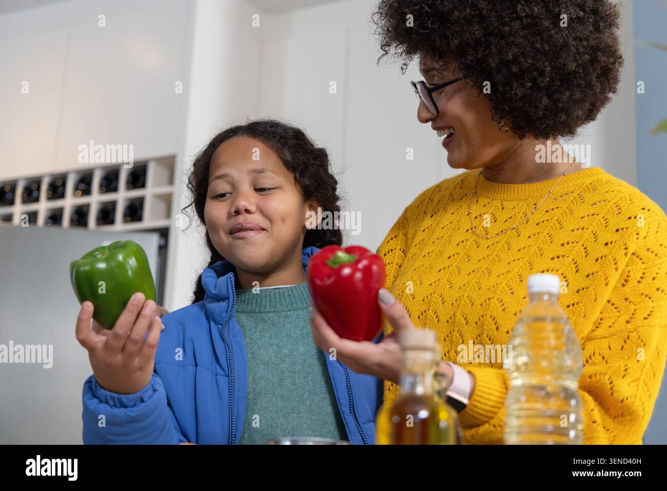 Verschiedene Mutter und Tochter halten grüne und rote Paprika in der Nähe der Ölflasche auf der Theke und untersuchen sie Stockfoto