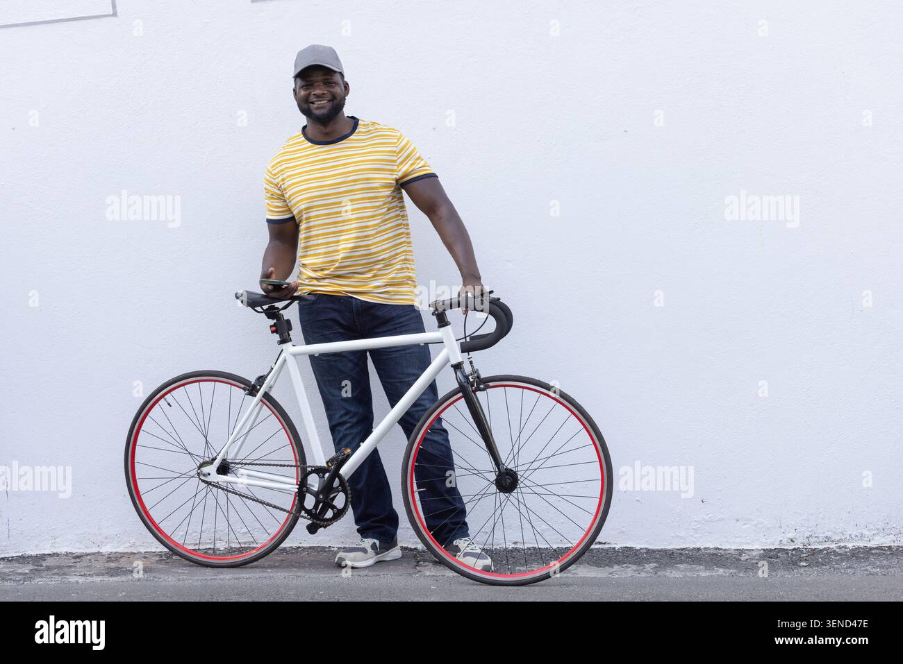 Erwachsener afrikanischer Mann, der sich auf ein Straßenfahrrad stützt und das Smartphone auf dem Bürgersteig gegen die weiße Wand hält Stockfoto