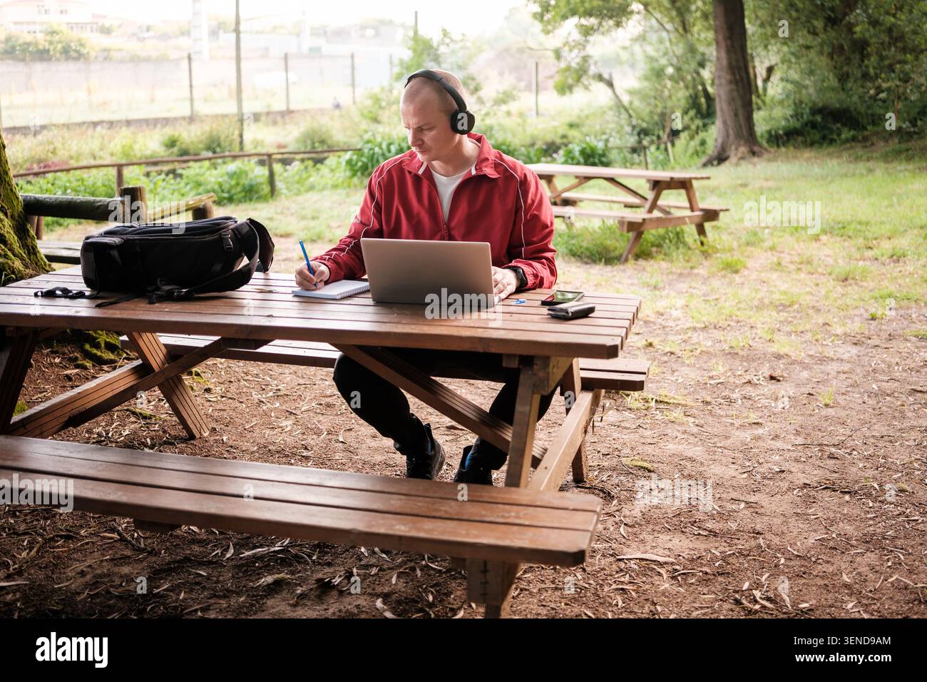 Mann, der Kopfhörer trägt und einen Laptop benutzt, während er an einem Picknicktisch im Park auf ein Notebook schreibt Stockfoto