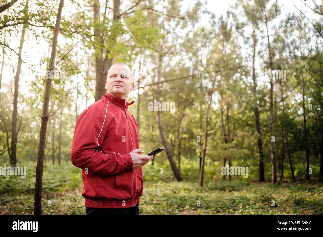 Mann in roter Jacke, der sich umsieht, Handy in einem Wald hält Stockfoto