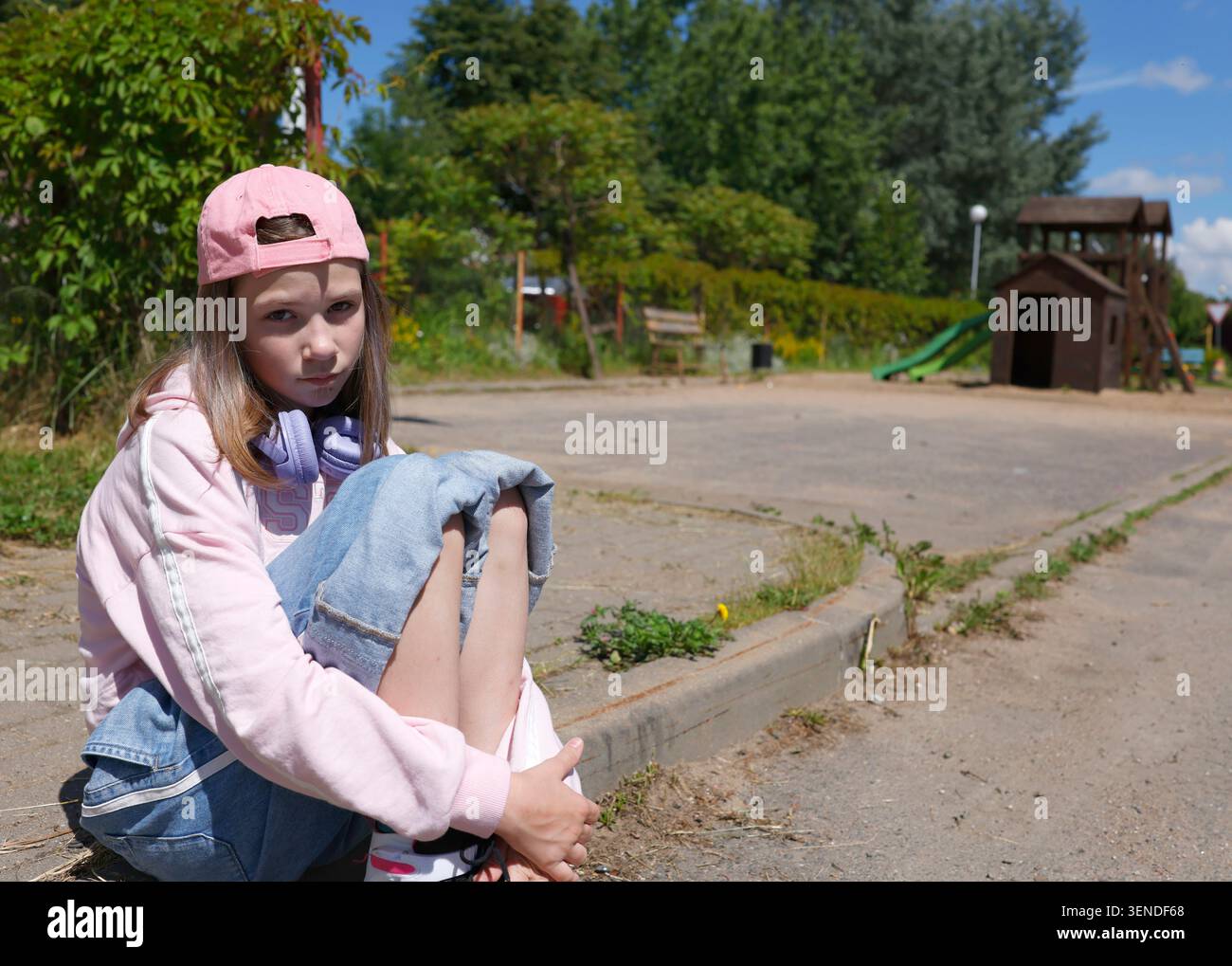 Trauriges Alpha-Mädchen der Generation, das auf einem Straßenrand sitzt und direkt in die Kamera blickt. Verärgertes Kind mit rosa Mütze und Jeans, die sich an die Knie schmiegt Stockfoto