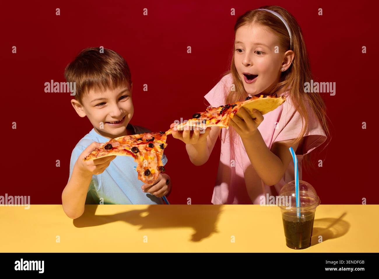 Zwei Kinder dehnen geschmolzenen Käse zwischen Pizzablättern auf rotem Studiohintergrund. Stockfoto