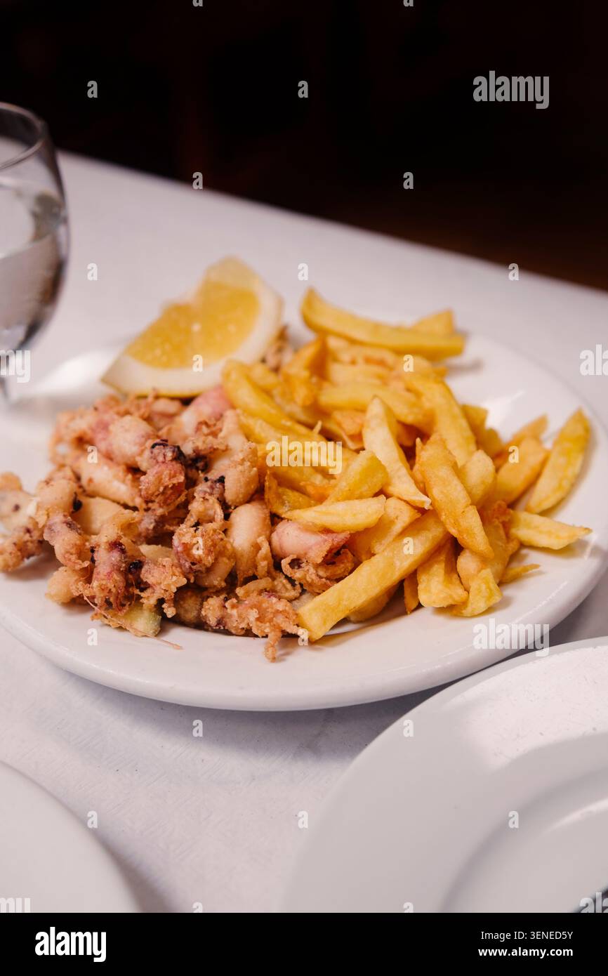 Knusprig gebratener Baby-Tintenfisch und Pommes frites serviert mit Zitronenkeil auf weißem Teller im spanischen Restaurant. Mediterrane Meeresfrüchte. Stockfoto