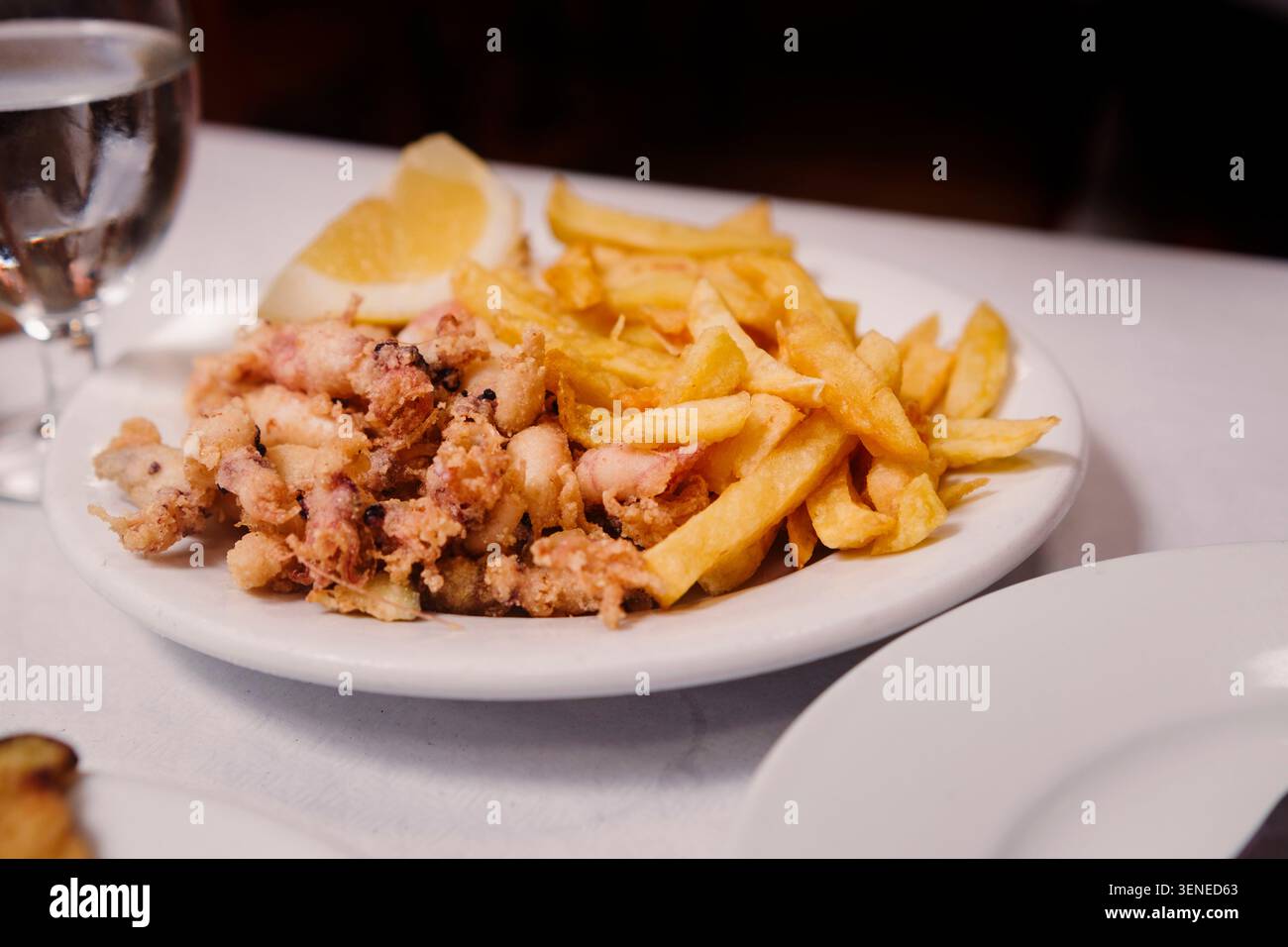 Knusprig gebratener Baby-Tintenfisch und Pommes frites serviert mit Zitronenkeil auf weißem Teller im spanischen Restaurant. Mediterrane Meeresfrüchte. Stockfoto